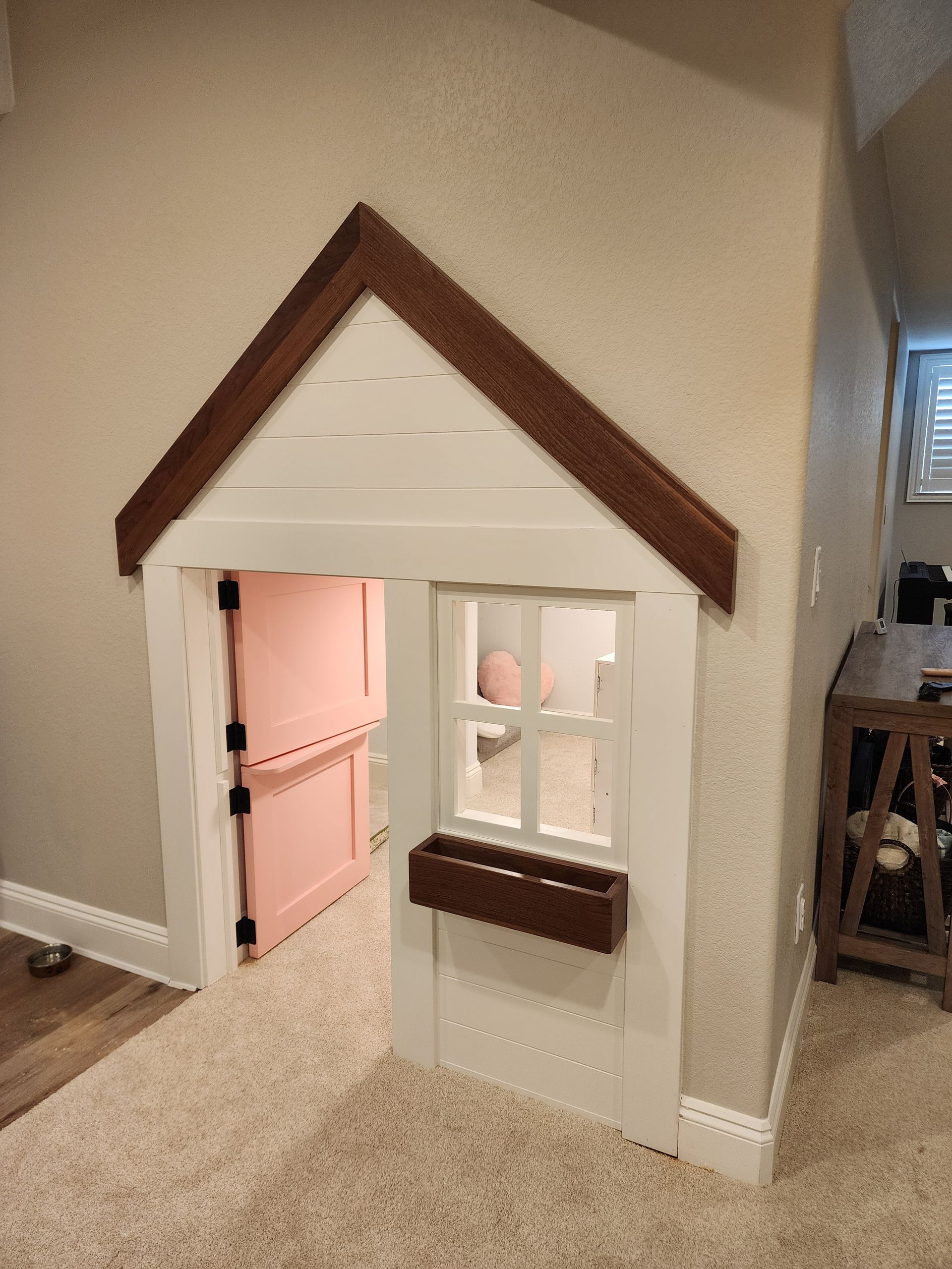 Playhouse built into a wall, white with a brown roof and a window. Door is pink.
