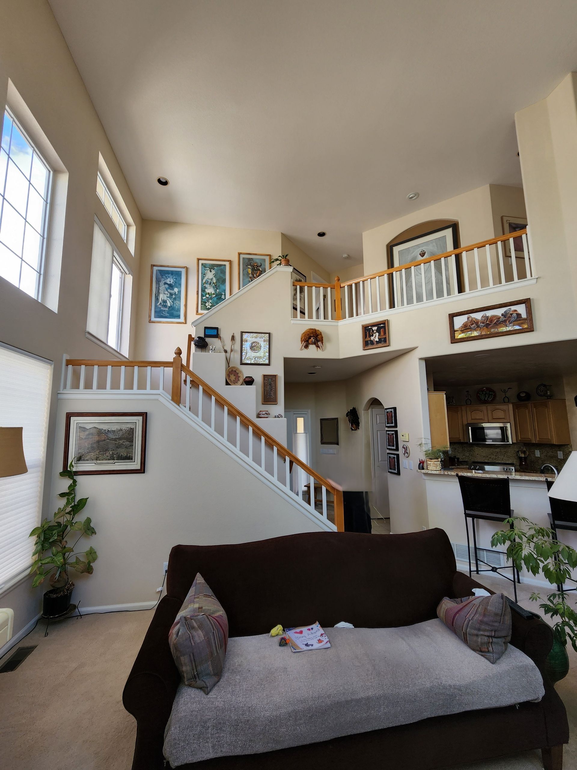 Open-plan living room with staircase, balcony, and dark brown sofa. Beige walls, wood accents, and multiple framed pictures.