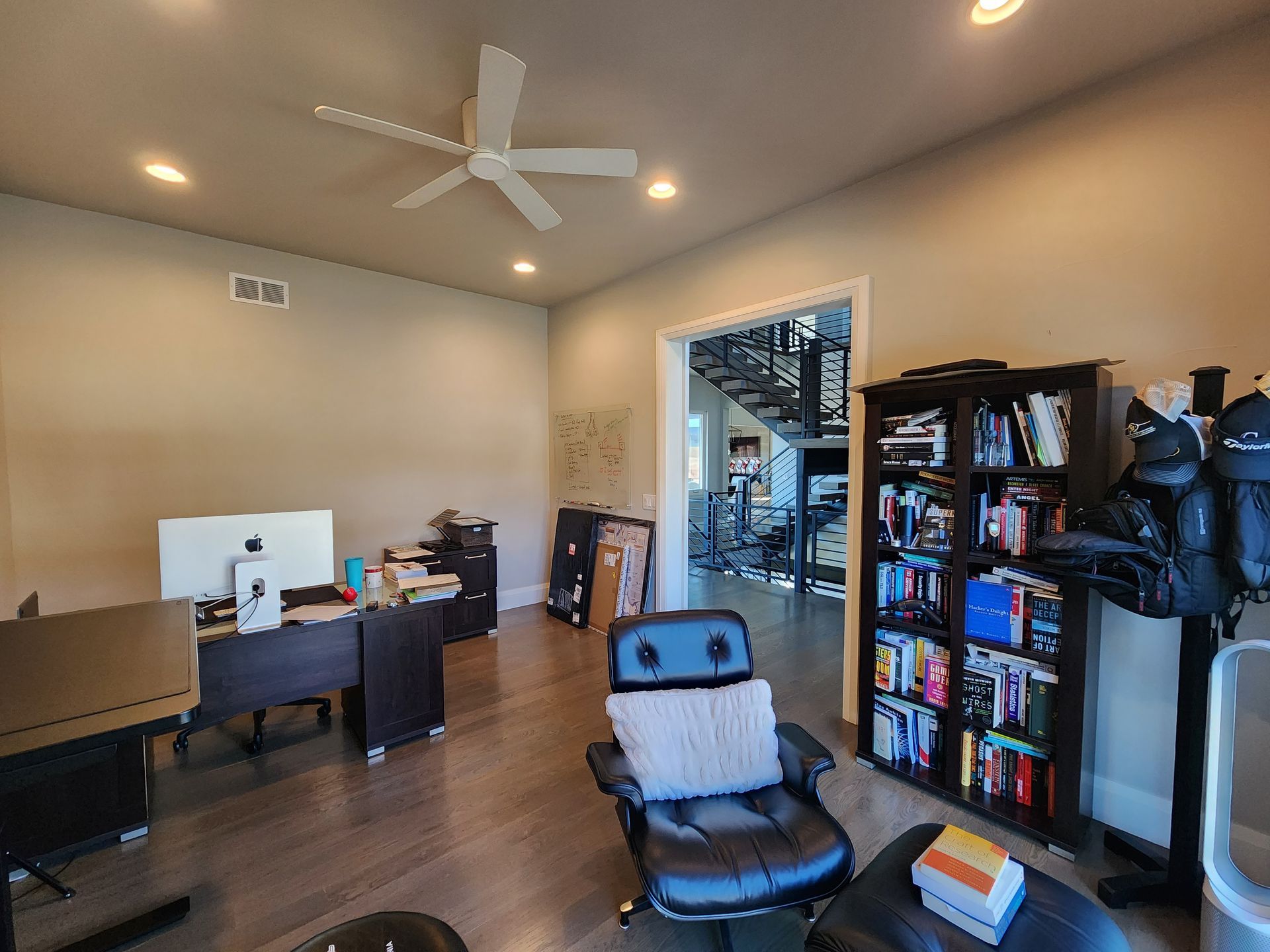 Office with desk, bookcase, and chair; doorway leads to stairs. Beige walls and hardwood floor.