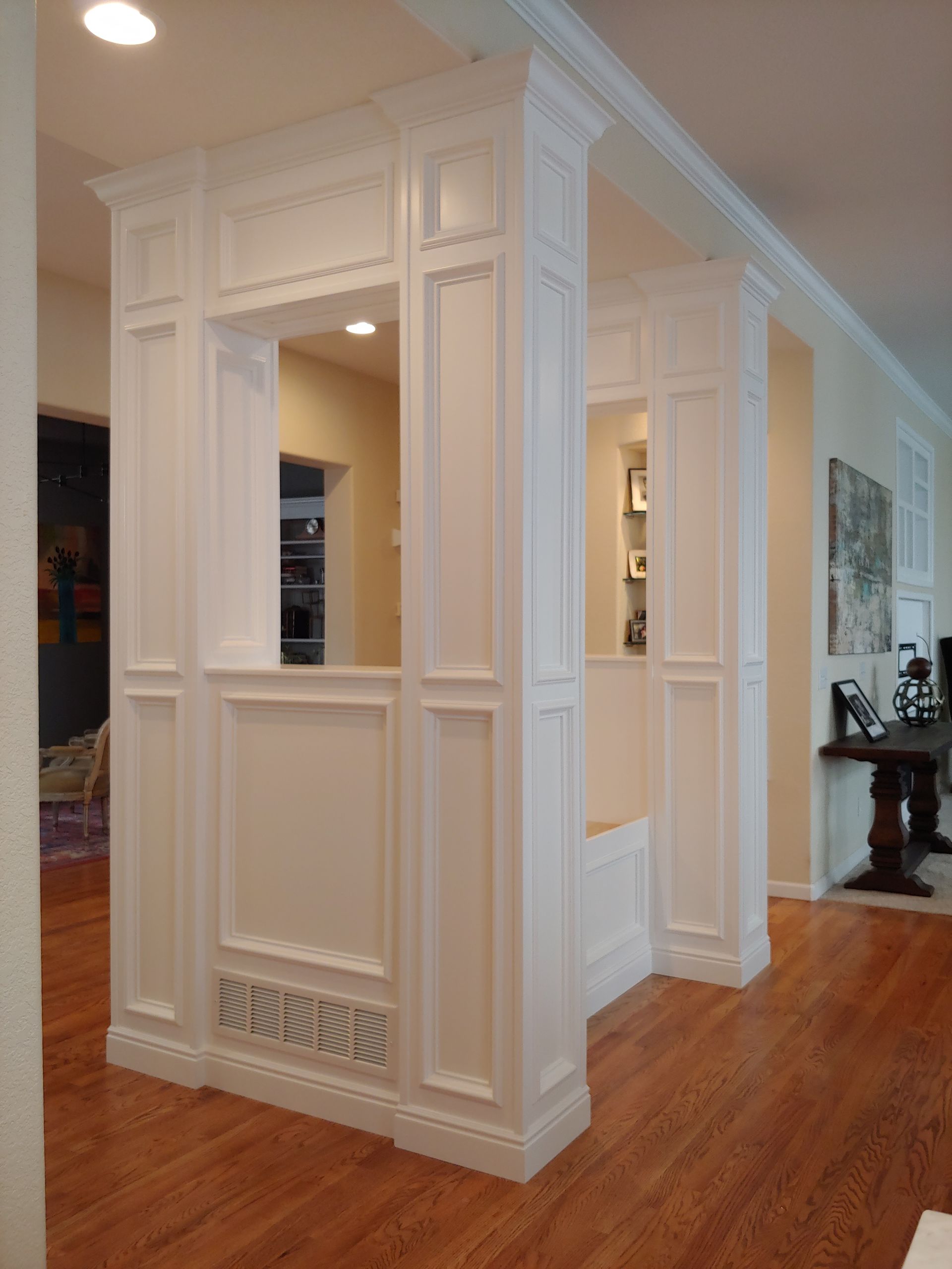 White-paneled architectural structure with a pass-through opening and built-in bench, set in a room with hardwood flooring.