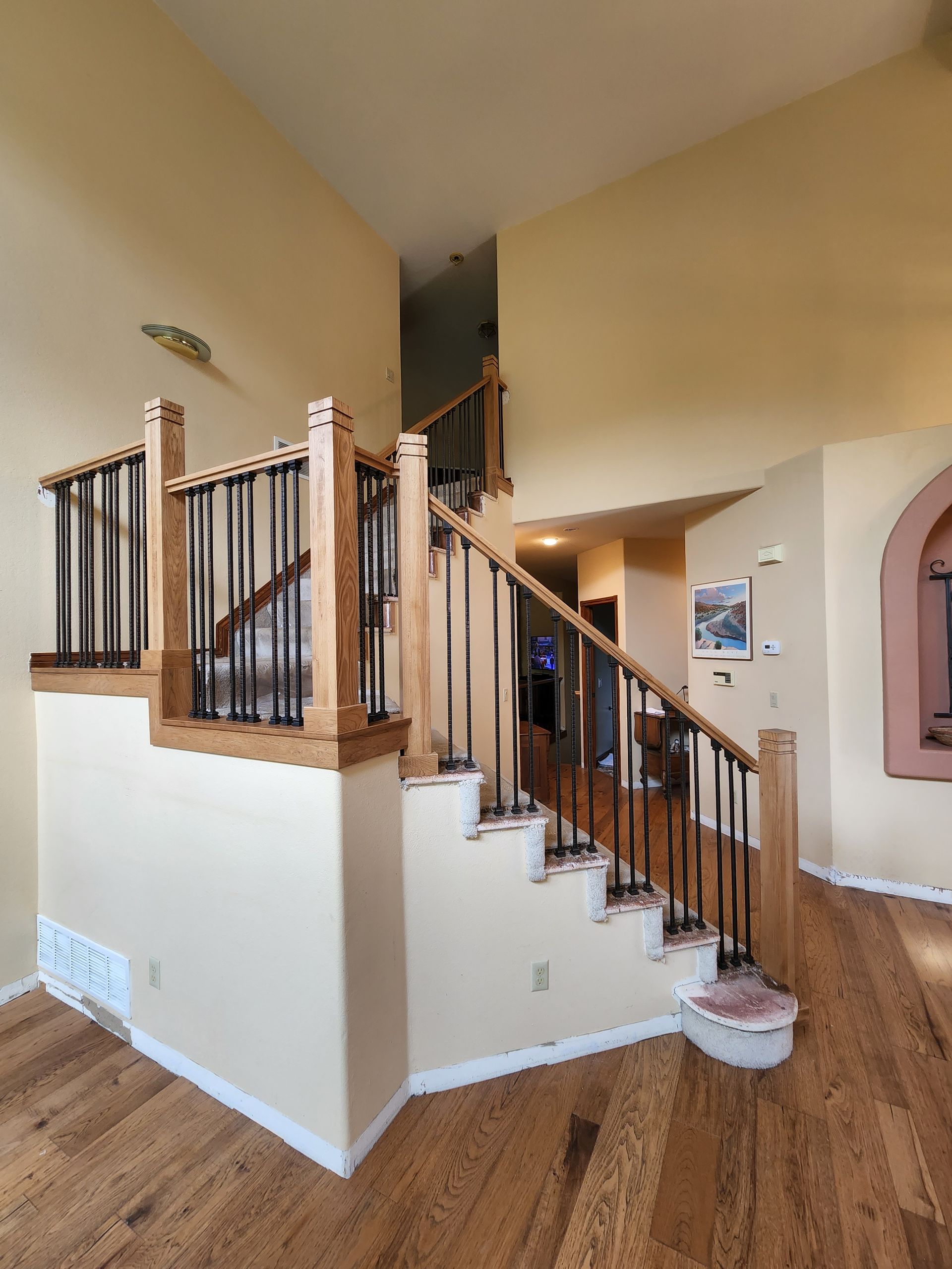 Staircase with wooden steps and railing, black iron balusters, hardwood floors, and tan walls.