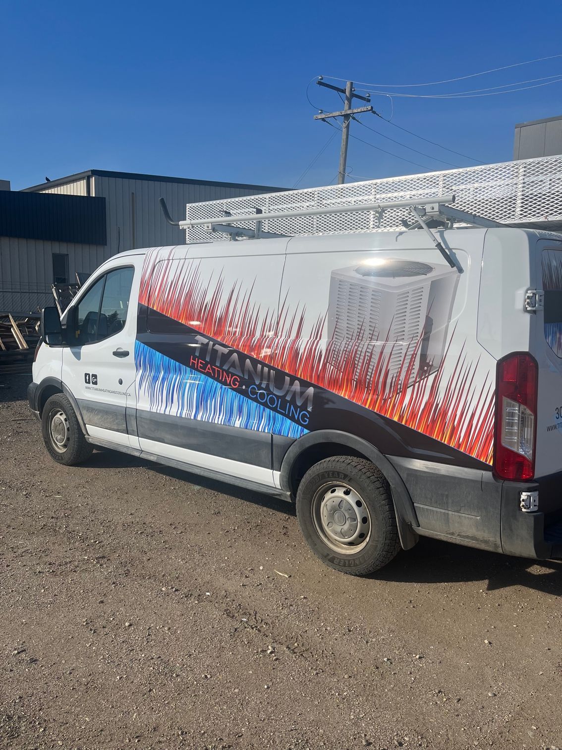 A white van with a ladder on top of it is parked in a gravel lot.