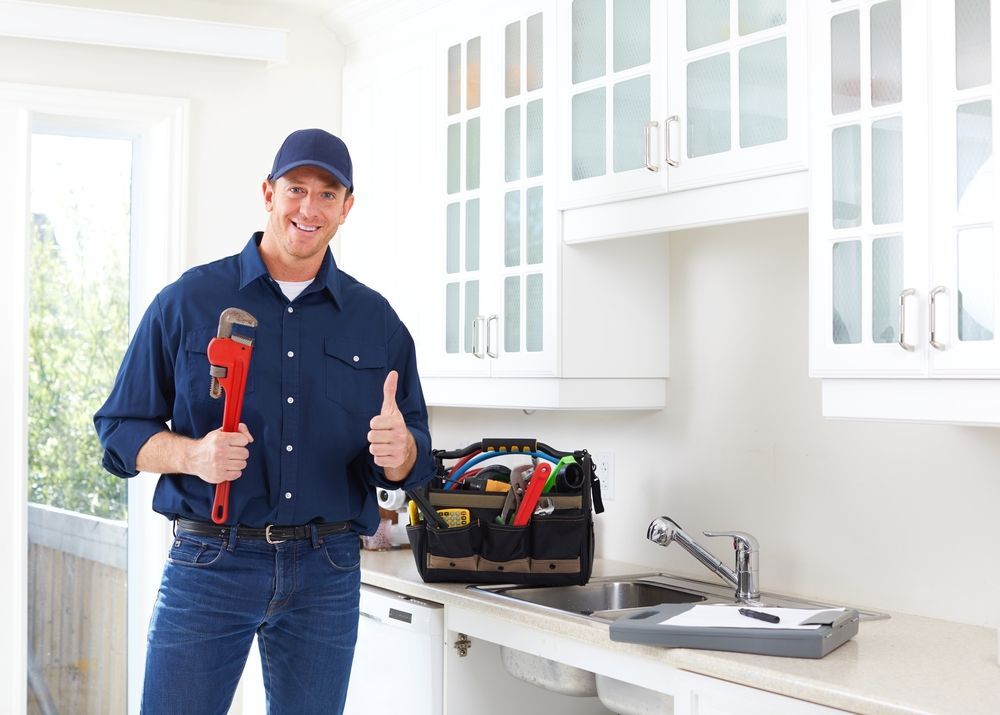 A plumber is standing in a kitchen holding a wrench and giving a thumbs up.