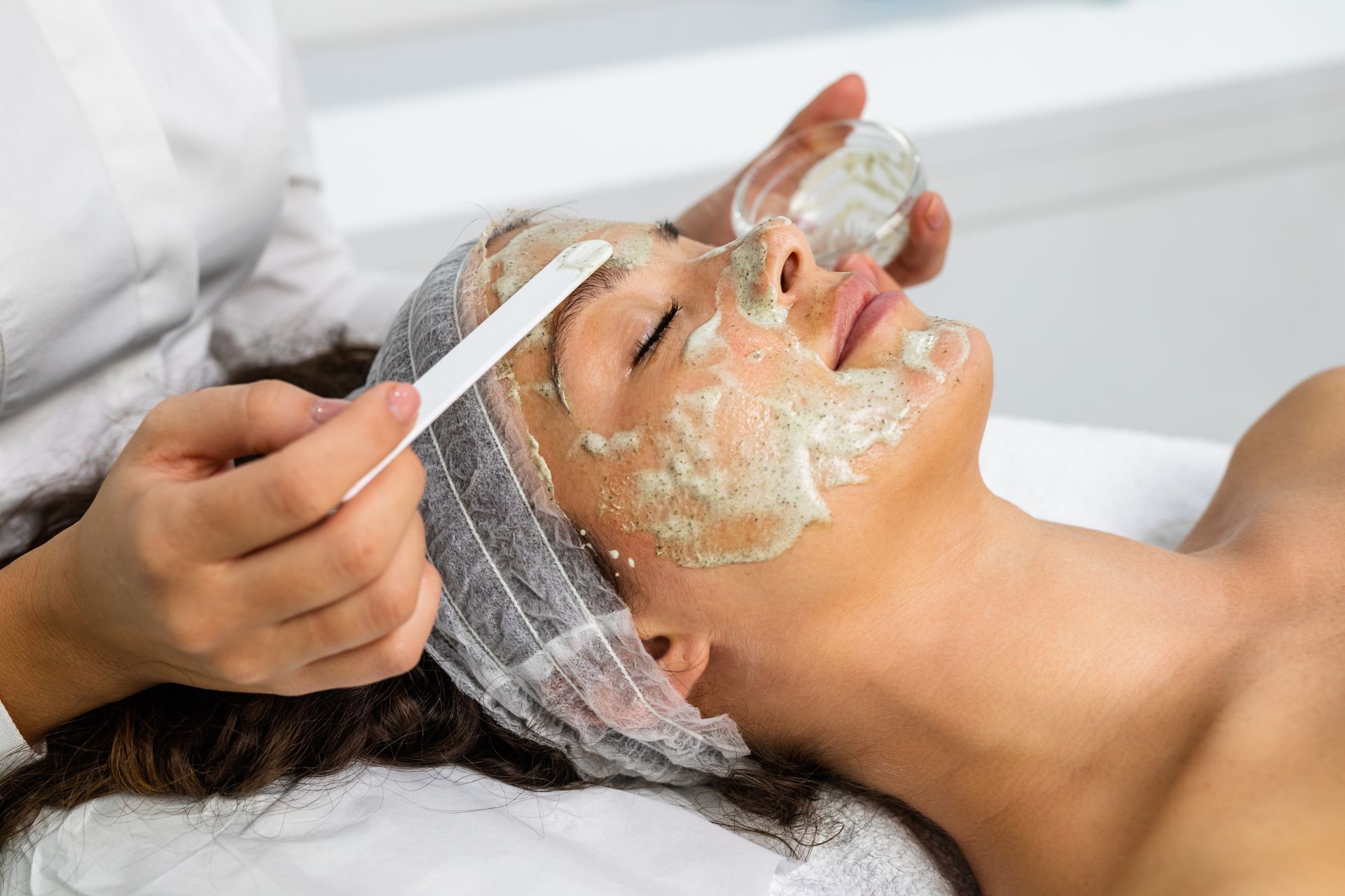 Woman receiving a facial mask in a spa; esthetician applying mask with a spatula.