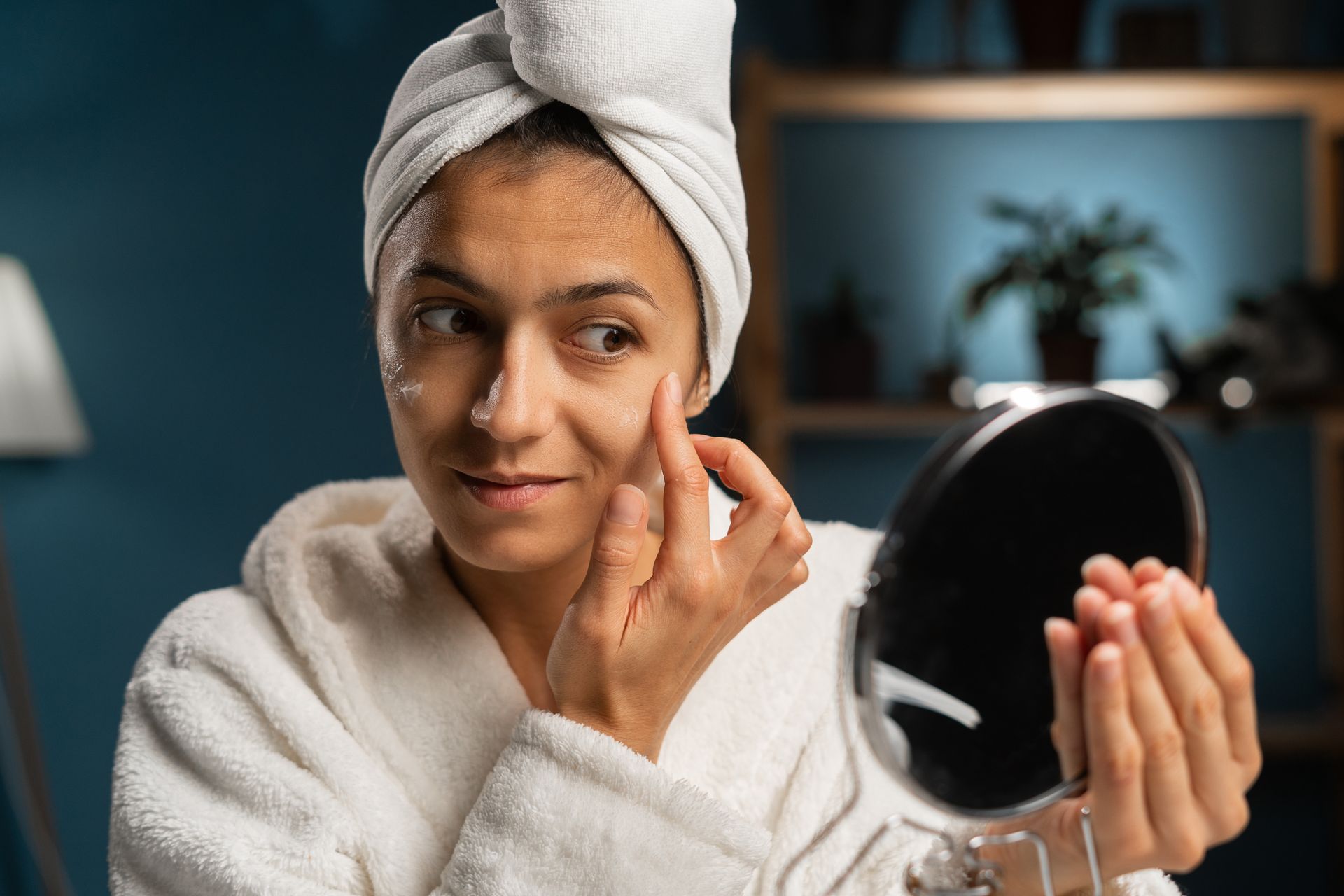Woman in a white robe applying makeup while looking into a handheld mirror in a softly lit room