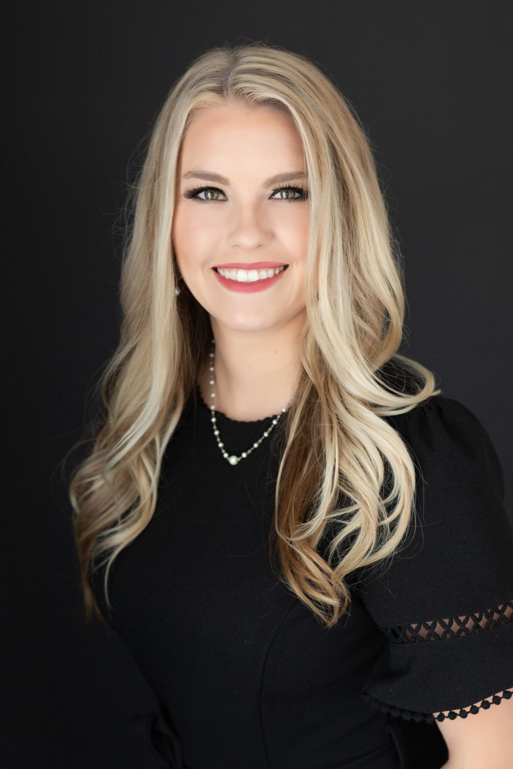 A woman in a black dress and necklace is smiling for the camera.