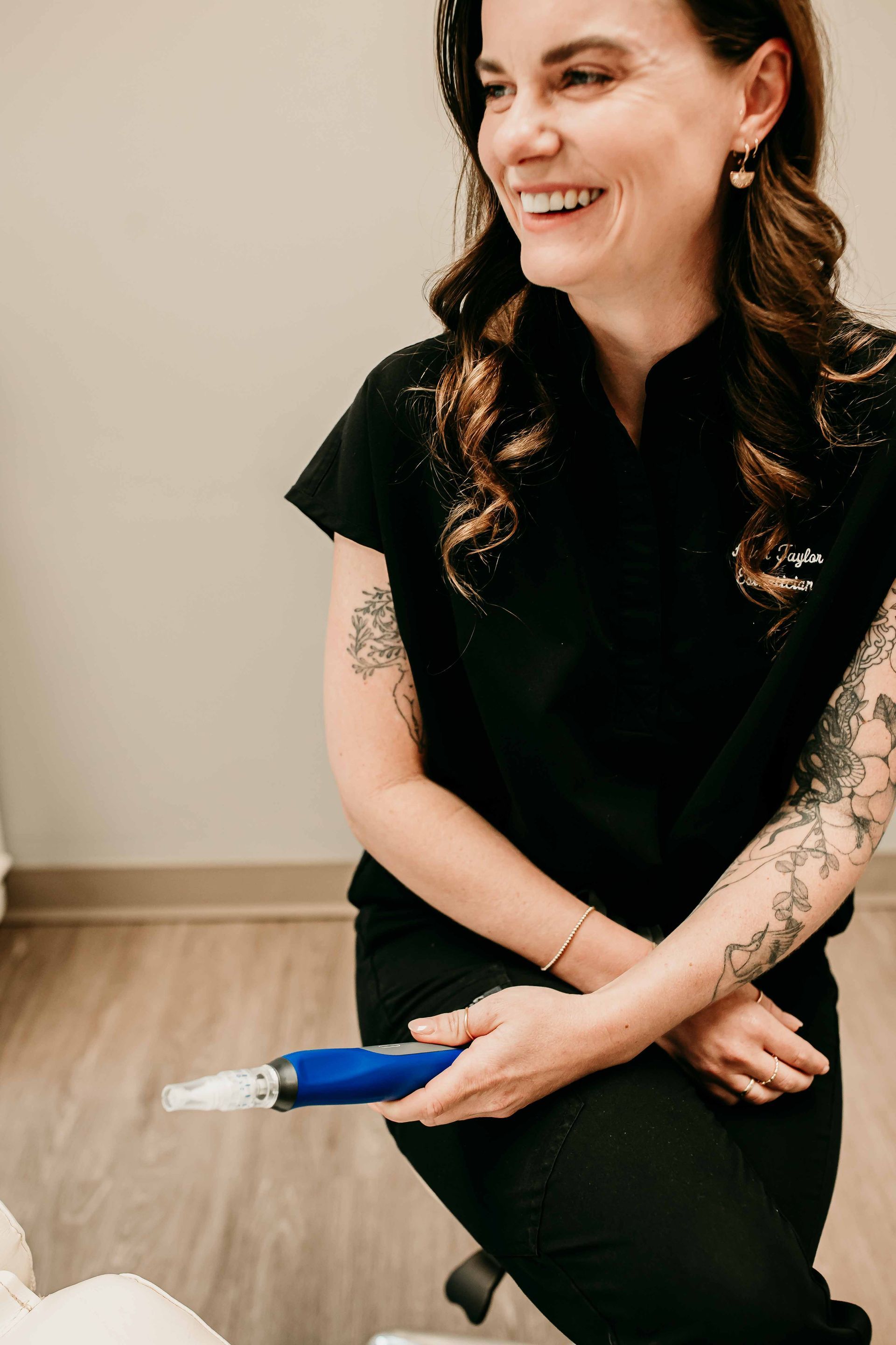 A professional in black scrubs holding a blue microneedling device, smiling in a clinical setting.