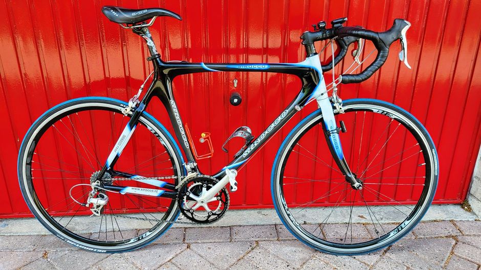 Racefiets met een zwart, blauw en wit frame tegen een rode deur.