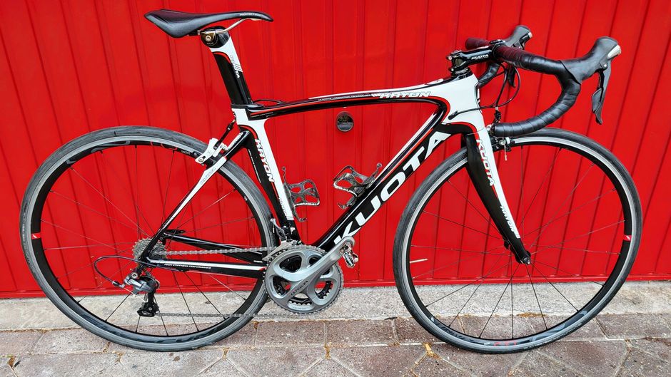 Zwart-witte Kuota racefiets met rode accenten tegen een rode deur.