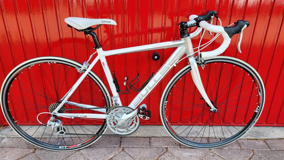 Witte racefiets met wit zadel, wit stuur en rode achtergrond.