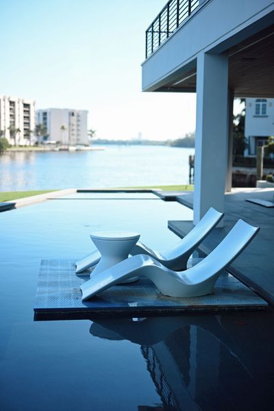 Infinity pool with two white lounge chairs beside a modern waterfront house.