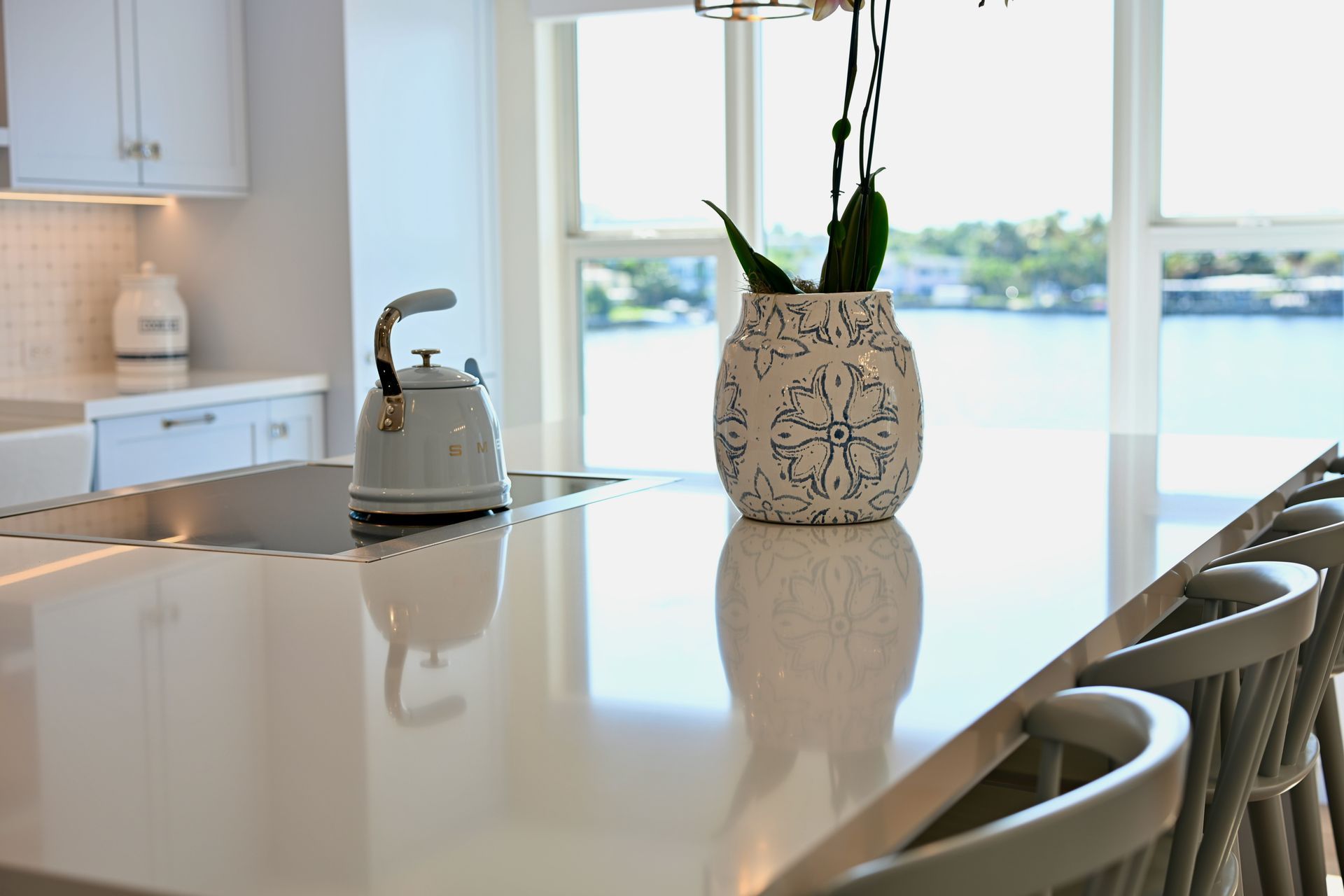 Bright kitchen island with a floral vase, kettle, and sunlight by a waterfront window
