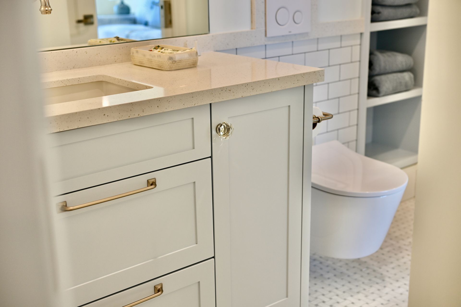 Light beige bathroom vanity with white countertop, brass hardware, and a small white trash bin beside open shelving.