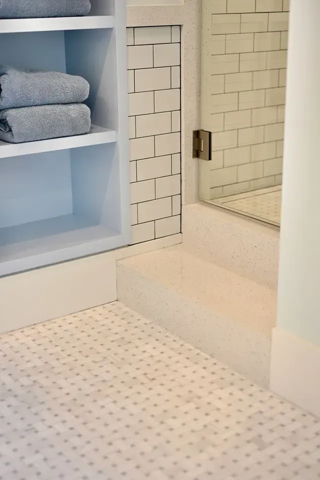 White-tiled bathroom with shower, glass door, built-in shelf, and folded towels on a blue wall shelf
