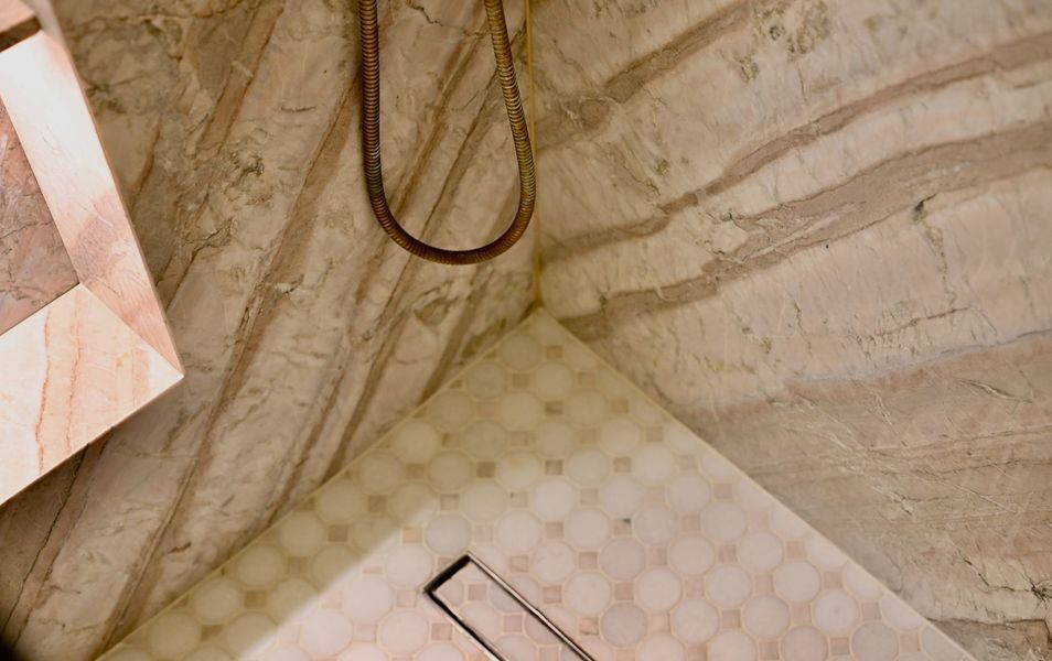 Bathroom corner with beige marble walls, shower hose, and a tiled floor drain cover