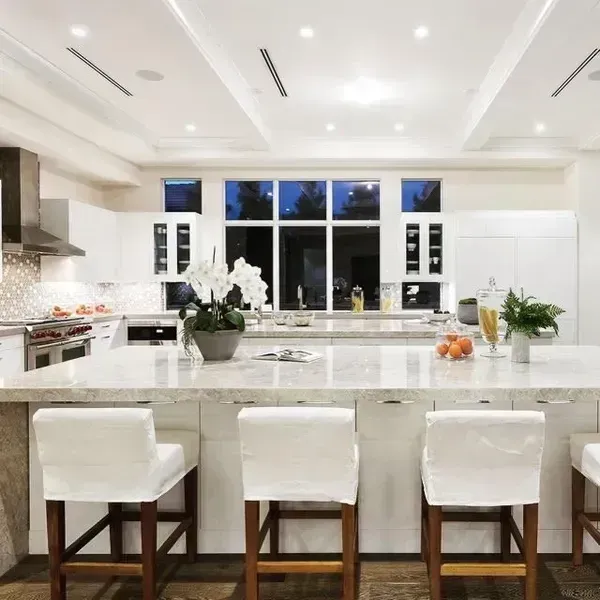 Bright modern white kitchen with marble island, four stools, flowers, and large windows at dusk