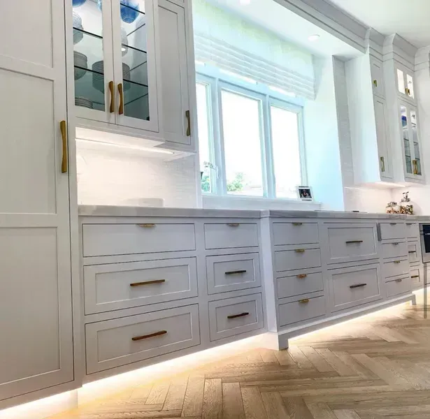 Bright white kitchen with built-in cabinets, marble countertop, and a large window over herringbone wood floor