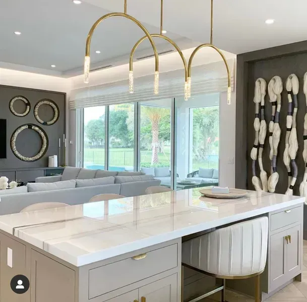 Modern open-concept kitchen with white island, gold pendant lights, and bright living area with large windows