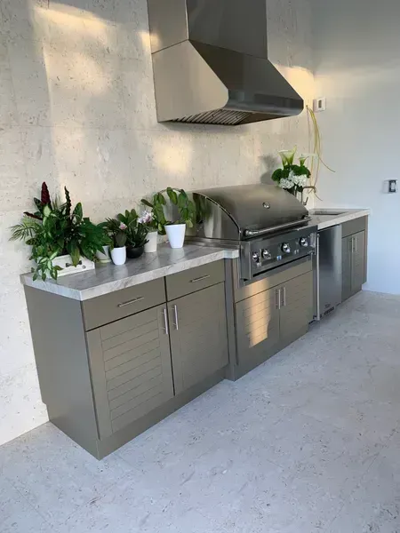 Modern outdoor kitchen with gray cabinets, stainless grill, hood, and potted plants on a stone patio.