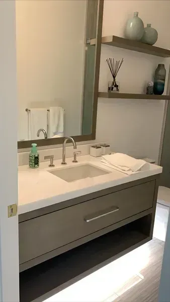 Modern bathroom vanity with white sink, mirror, shelves, and gray cabinet under soft lighting