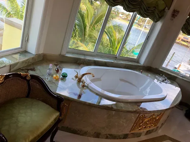 Elegant bathroom with a marble jacuzzi tub and ornate chair by large windows overlooking palm trees