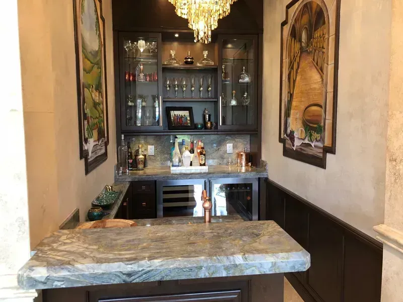 Narrow hallway bar with marble counter, dark cabinets, chandelier, and framed wall art.