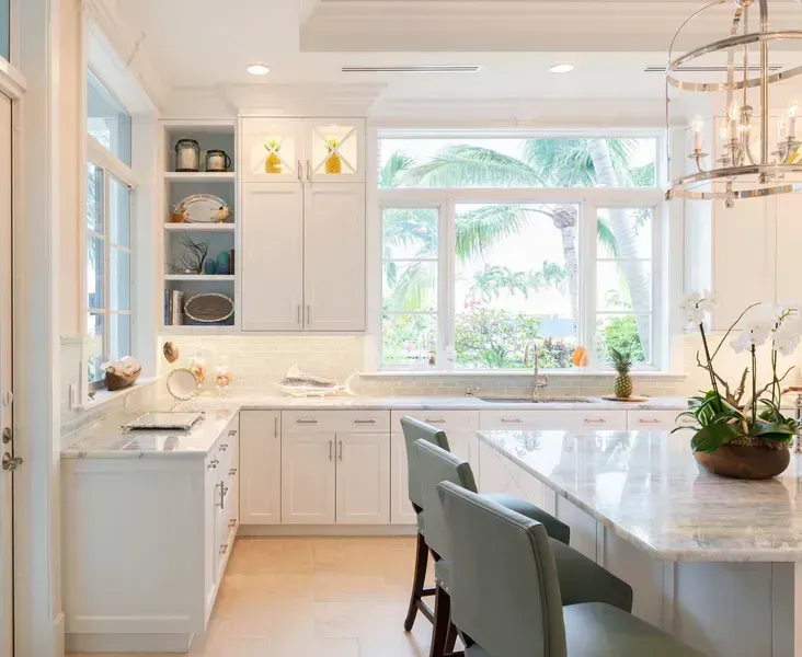 Bright modern kitchen with white cabinets, marble island, and large sunlit window overlooking palm trees