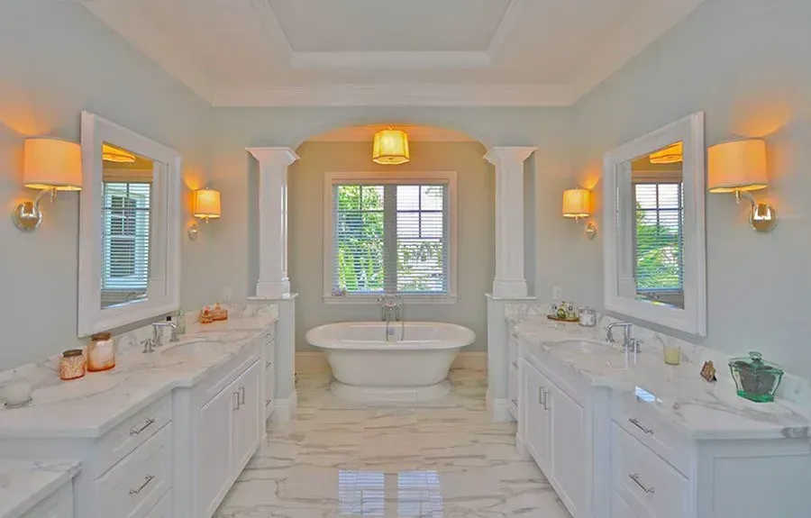 Bright white bathroom with double vanities, wall sconces, central tub, and a windowed alcove.