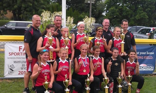 Team of children in red uniforms holding trophies on a podium outdoors, with adults behind them
