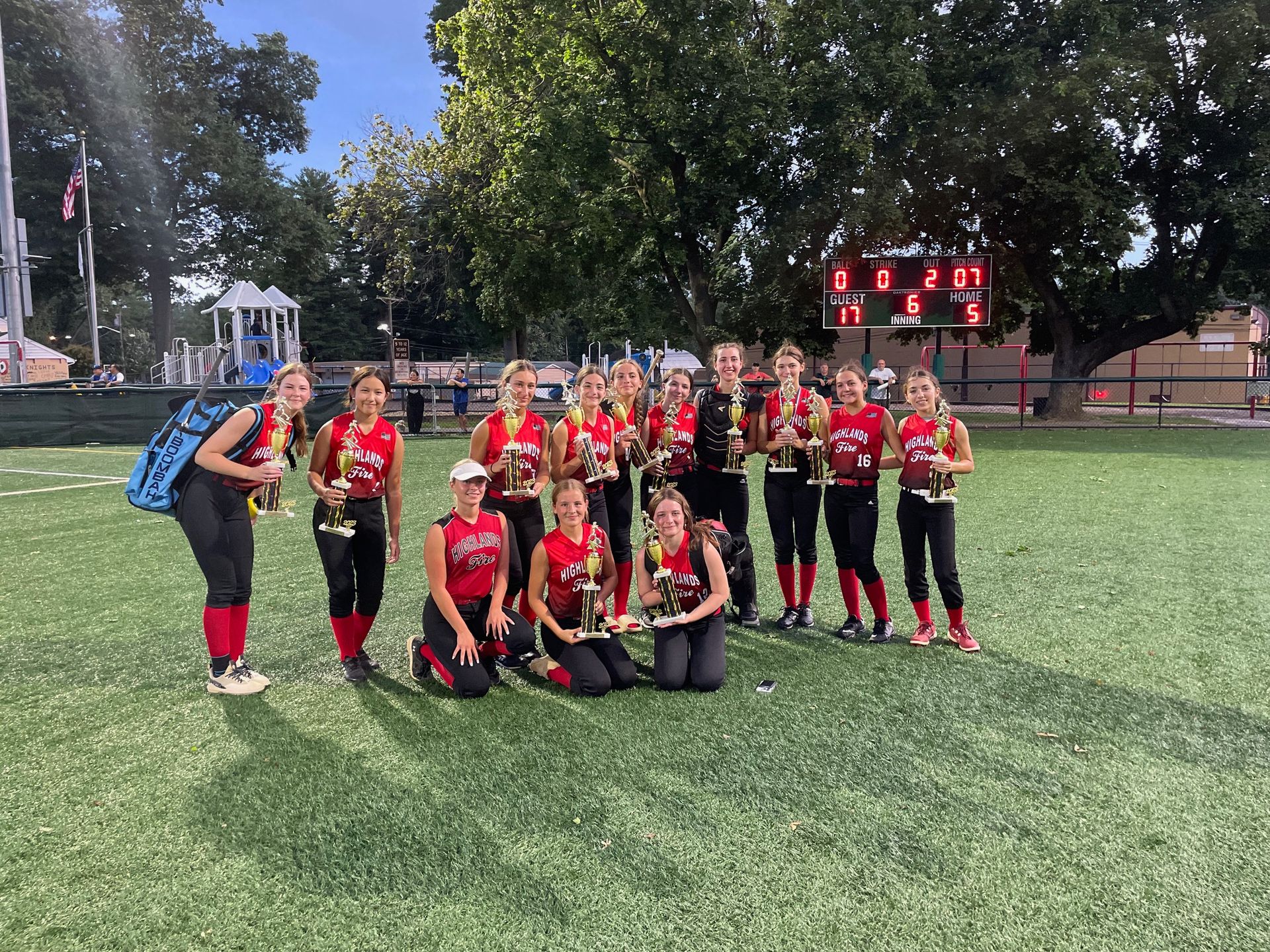 Youth sports team in red and black uniforms posing on a grassy field at dusk with a lit scoreboard behind them