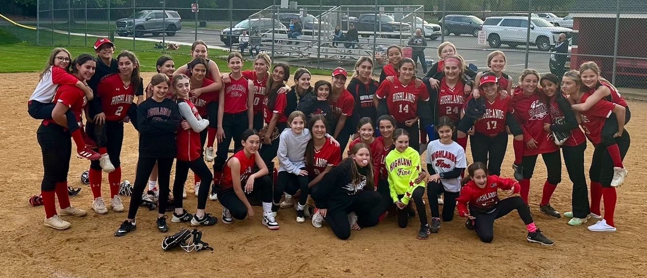 Youth softball team in red uniforms posing together on a dirt field, with a chain-link fence and parked cars behind them