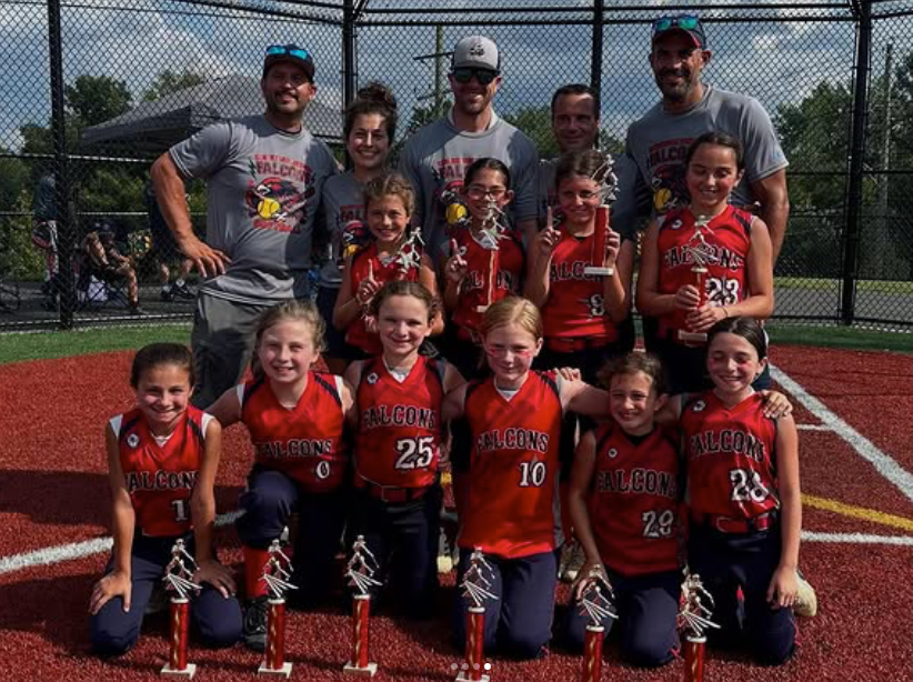 Youth softball team posing on a field with trophies, coaches standing behind them.