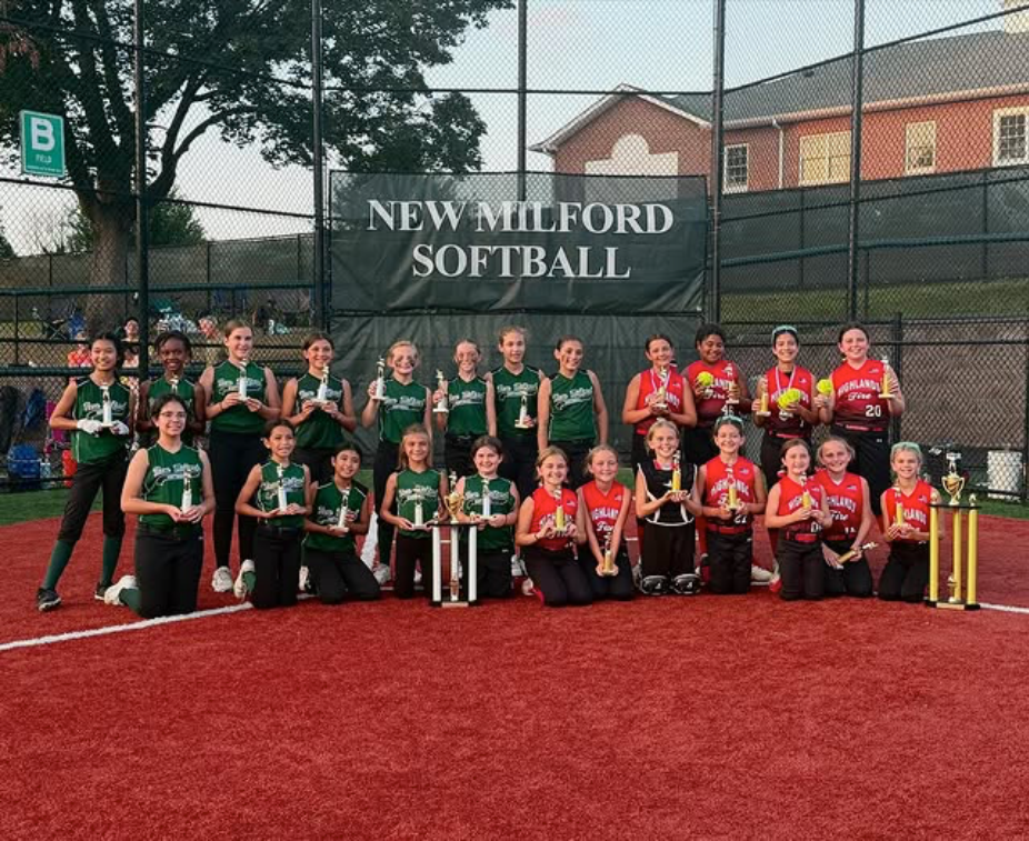 New Milford softball team posing with trophies on a red field in front of a chain-link fence