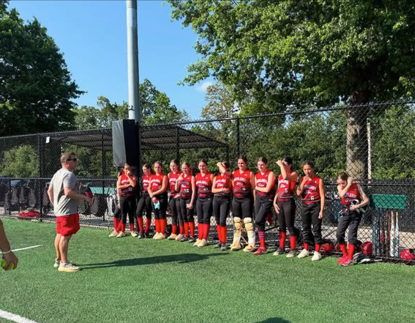 Youth sports team in red-and-black uniforms lined up on turf beside a coach, near a fenced field in sunshine