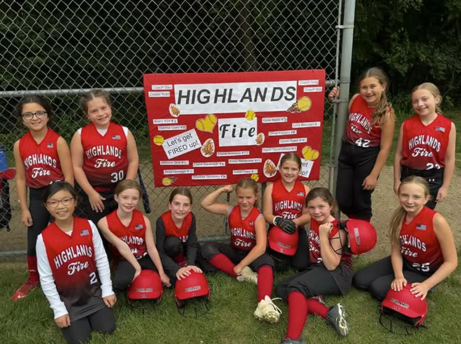 Youth baseball team posing in red uniforms with a “Highlands Fire” sign on a field.