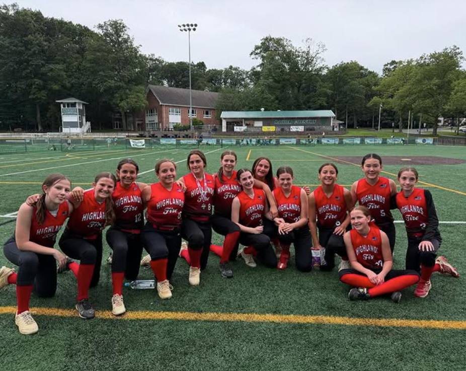Softball team in red uniforms posing on a field, kneeling and smiling before a game