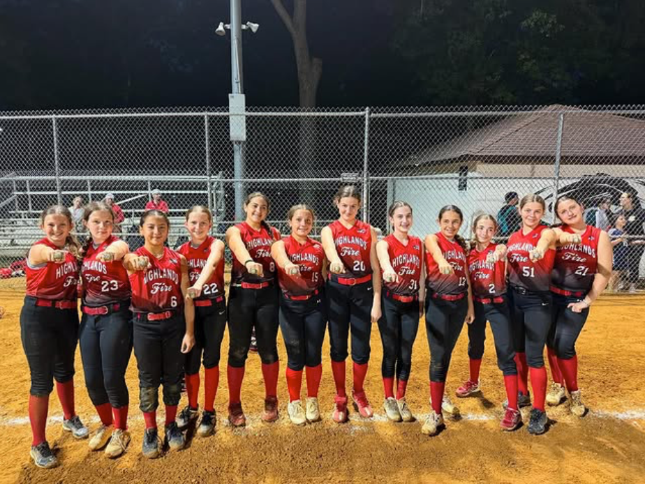 Softball team in red jerseys posing together on a field at night, holding gloves and smiling.