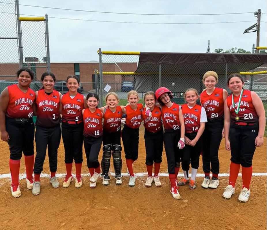 Youth softball team in red jerseys posing on a dirt field, holding bats and gloves, with a chain-link fence behind them