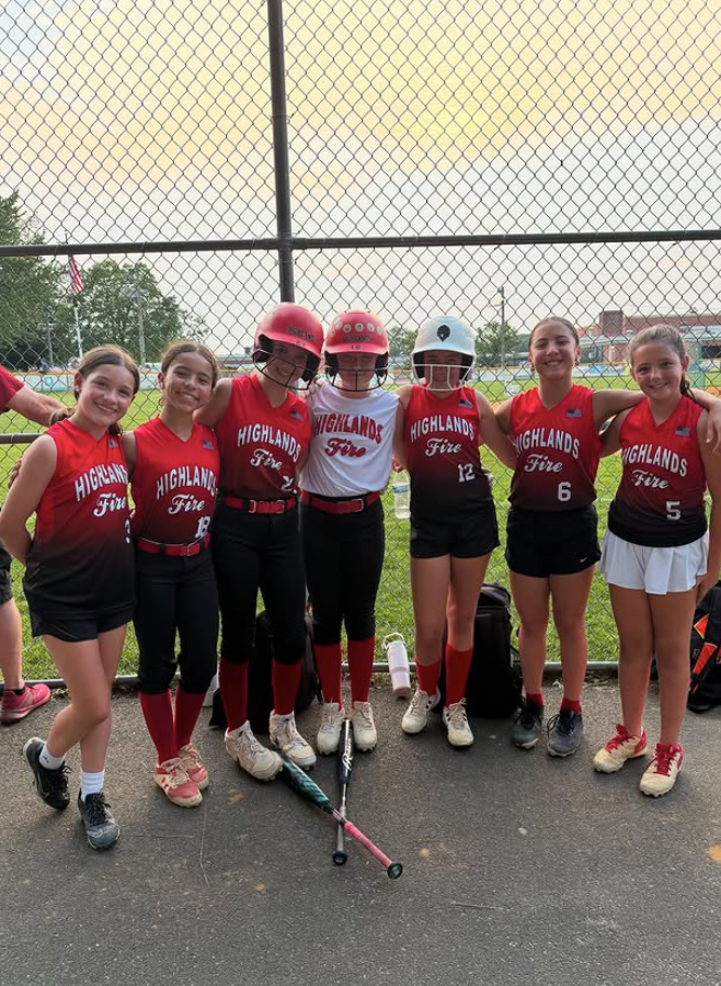 Youth field hockey team in red uniforms posing with sticks on an outdoor court, smiling in front of a fence.