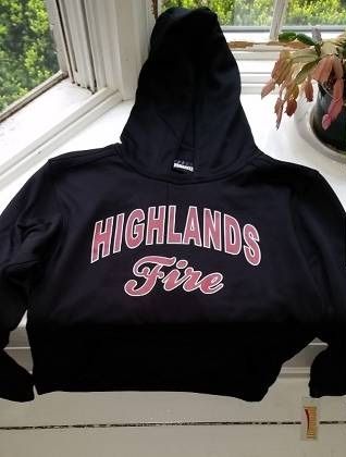 Black Highlands Travel Softball hoodie with “HIGHLANDS Fire” printed in pink and white, laid on a windowsill.