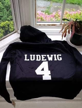 Black Highlands Travel Softball sports jersey with player name and number draped on a chair by a window