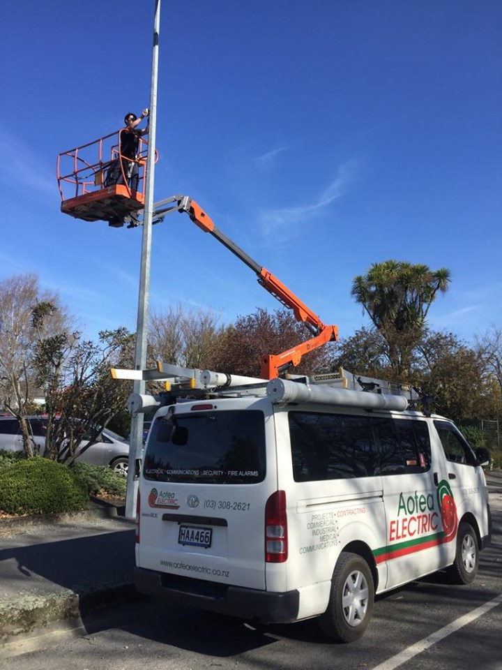 An electrician in a lift bucket works on a light pole; white van with 