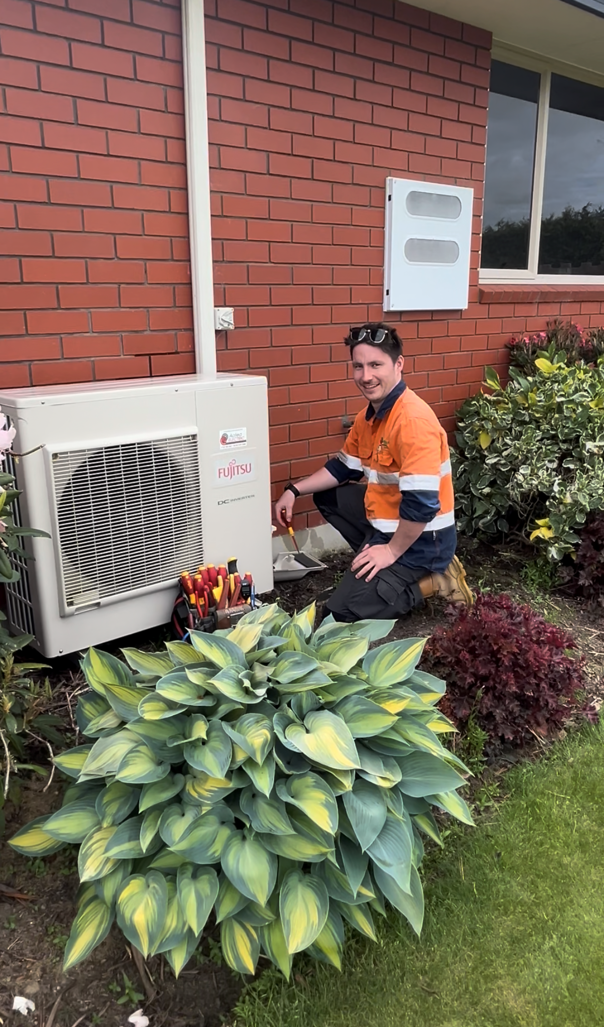 A repair technician in tan shirt and hat works on an outdoor HVAC unit, tool kit in front of them.