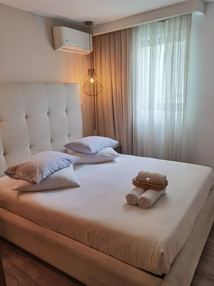 Cozy bedroom with tufted white bed, beige bedding, hanging lamp, air conditioner, and sheer curtains