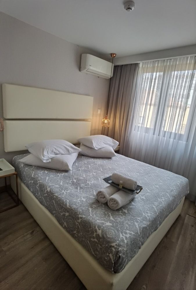 Bright bedroom with a double bed, gray bedding, white pillows, and sheer curtains by a window.