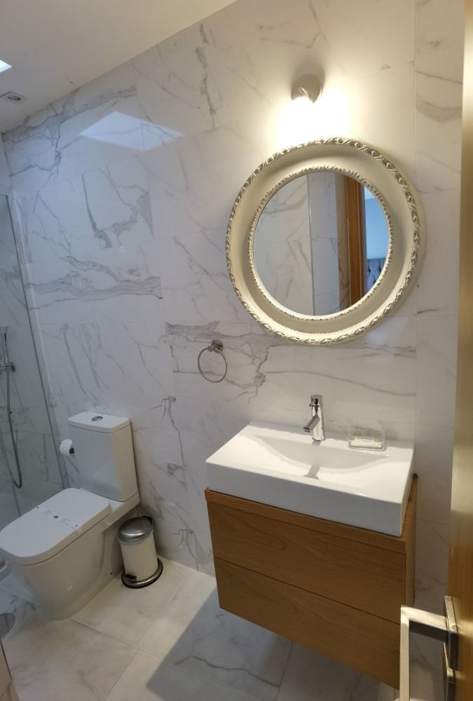 Small modern bathroom with marble-look walls, wood vanity, round mirror, toilet, and trash can.