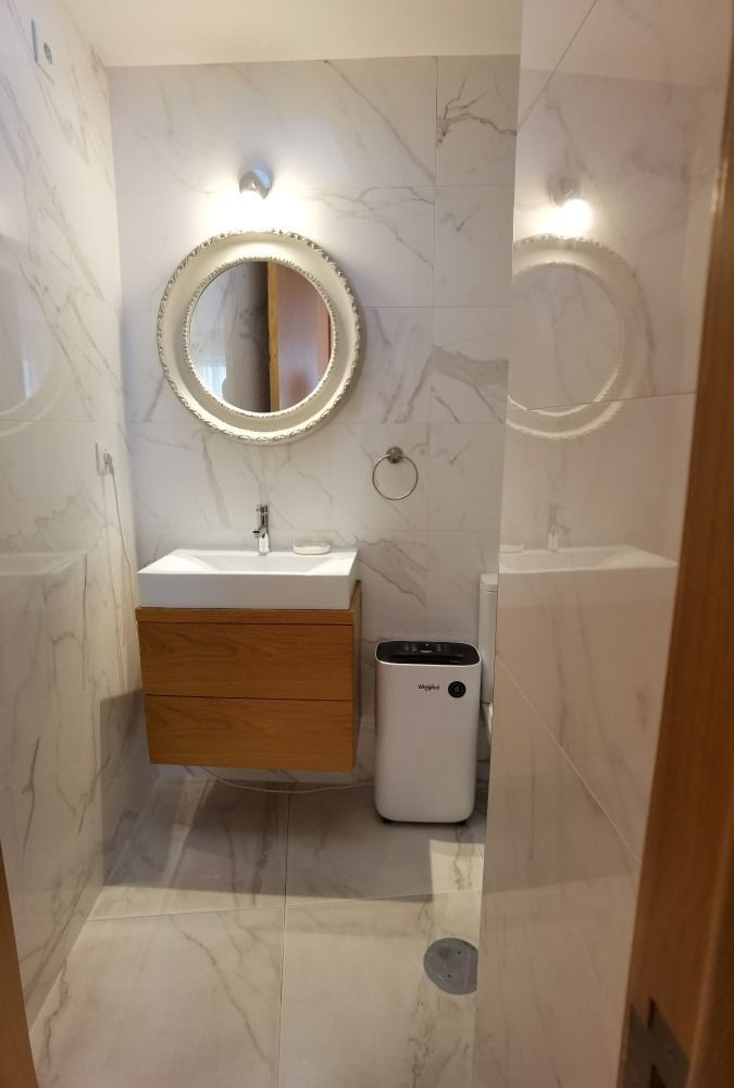 Modern bathroom with marble walls, round lit mirror, floating sink, and a toilet beside a shower drain