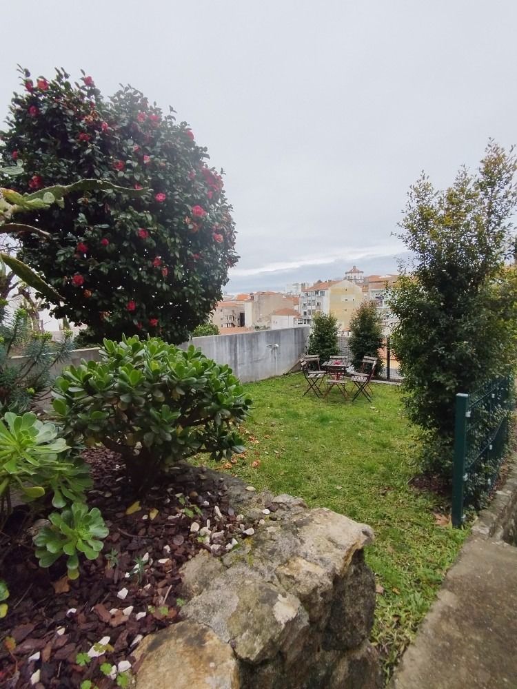 Garden with flowering shrubs, stone wall, and a grassy yard overlooking houses under a cloudy sky