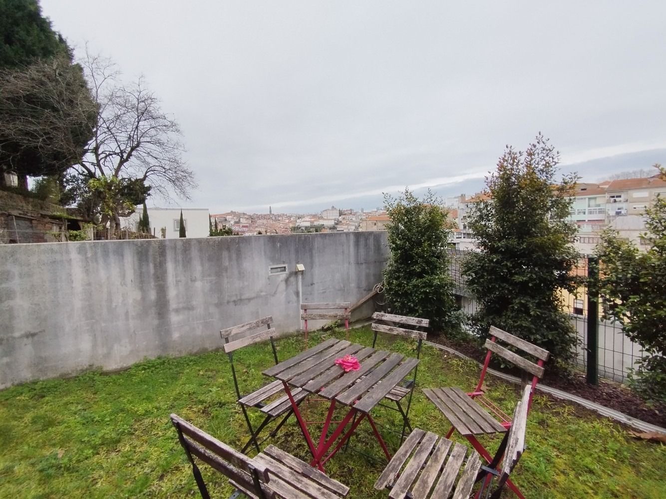 Tilted wooden table and chairs on a grassy yard beside a concrete wall under a cloudy sky
