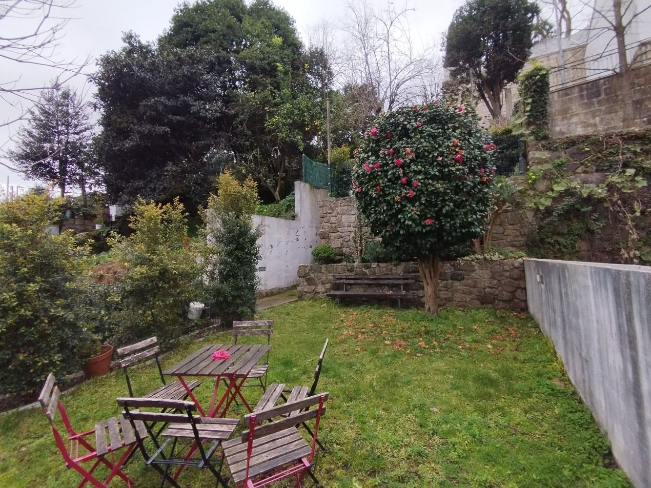 Small backyard garden with patio table and chairs, grassy lawn, stone steps, and flowering shrubs.